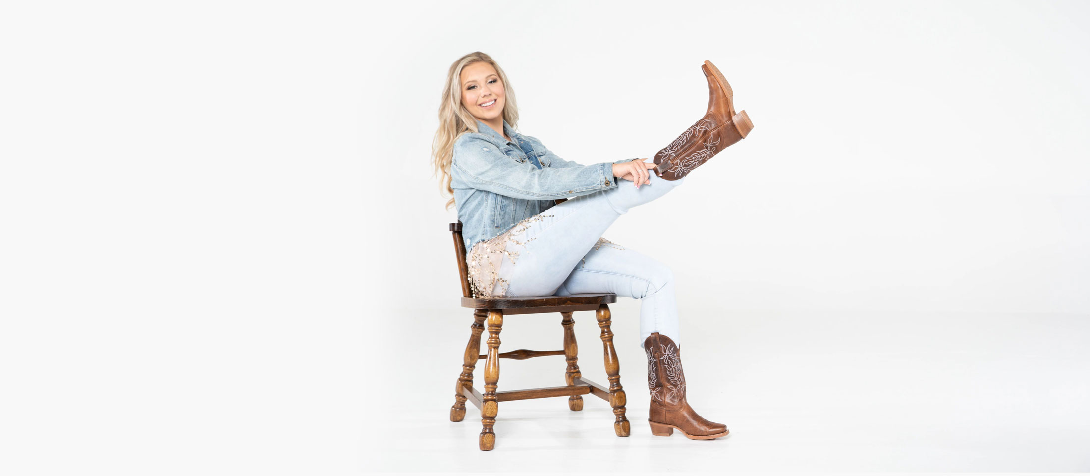 A woman wearing a jean jacket and jeans, sitting in a chair putting on cowboy boots.